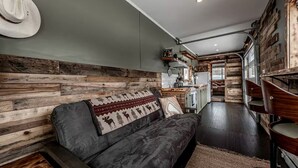 A cozy living area with a sofa, wooden wall paneling, and a kitchen in the background. - Serene Recycled Container in Big Sky Montana, Perfect for a Glamping Weekend in the Wilderness (Ovando)