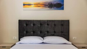 Un lit avec une tête de lit capitonnée, deux oreillers et un tableau représentant un coucher de soleil sur l’eau.