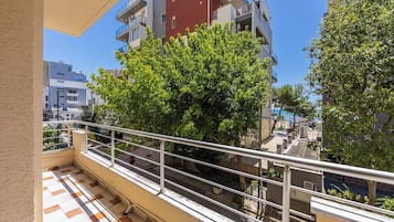 En balkon med gelænder, flisegulv og udsigt til et træ og bygninger.