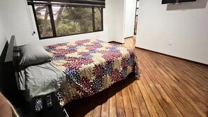 Ein Schlafzimmer mit einem Bett, einem Fenster mit Jalousien, einem an der Wand befestigten Fernseher und einem Holzboden.