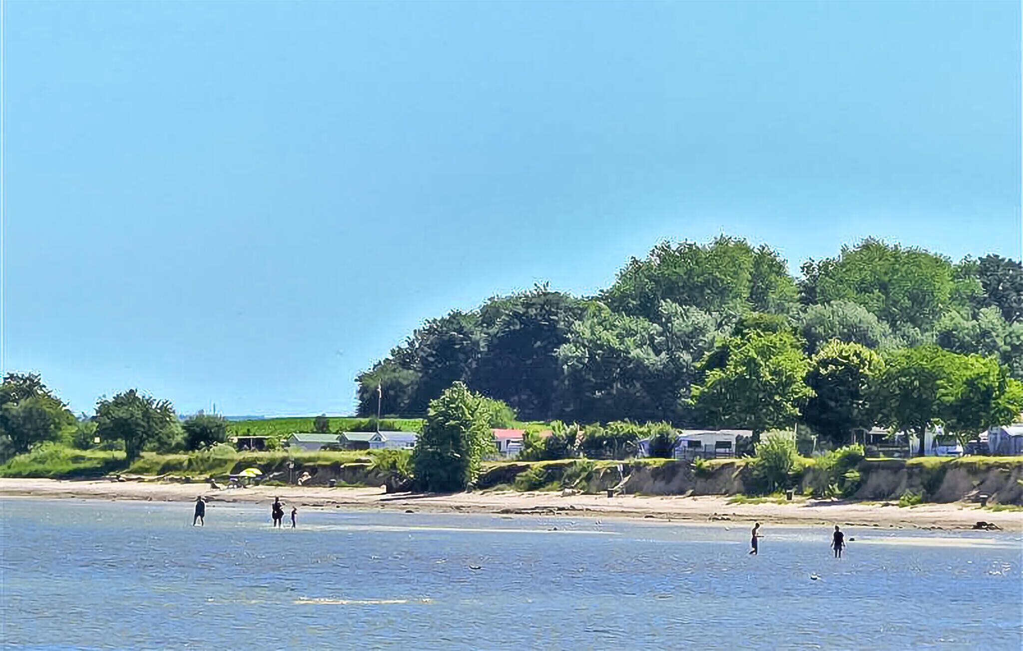 Une plage où des gens se promènent le long du rivage, un ciel dégagé et une colline avec des arbres en arrière-plan.