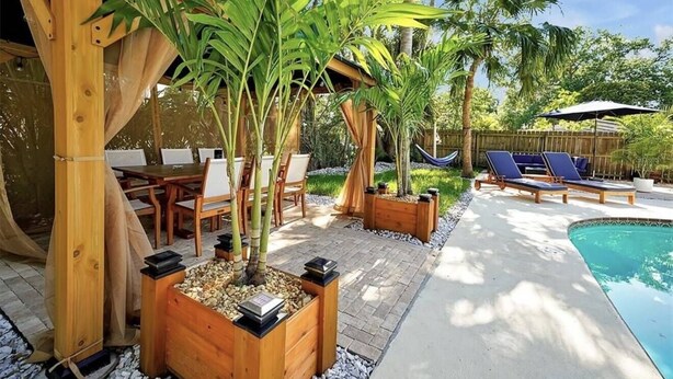 Una pérgola de madera, mobiliario exterior, una piscina y plantas en macetas.