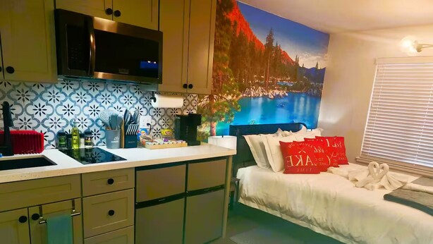 Une cuisine moderne équipée d’un four encastré, d’un four à micro-ondes et d’un évier. On y trouve un lit avec une literie blanche et des oreillers rouges. Un grand tableau représentant un paysage montagneux est accroché au mur.