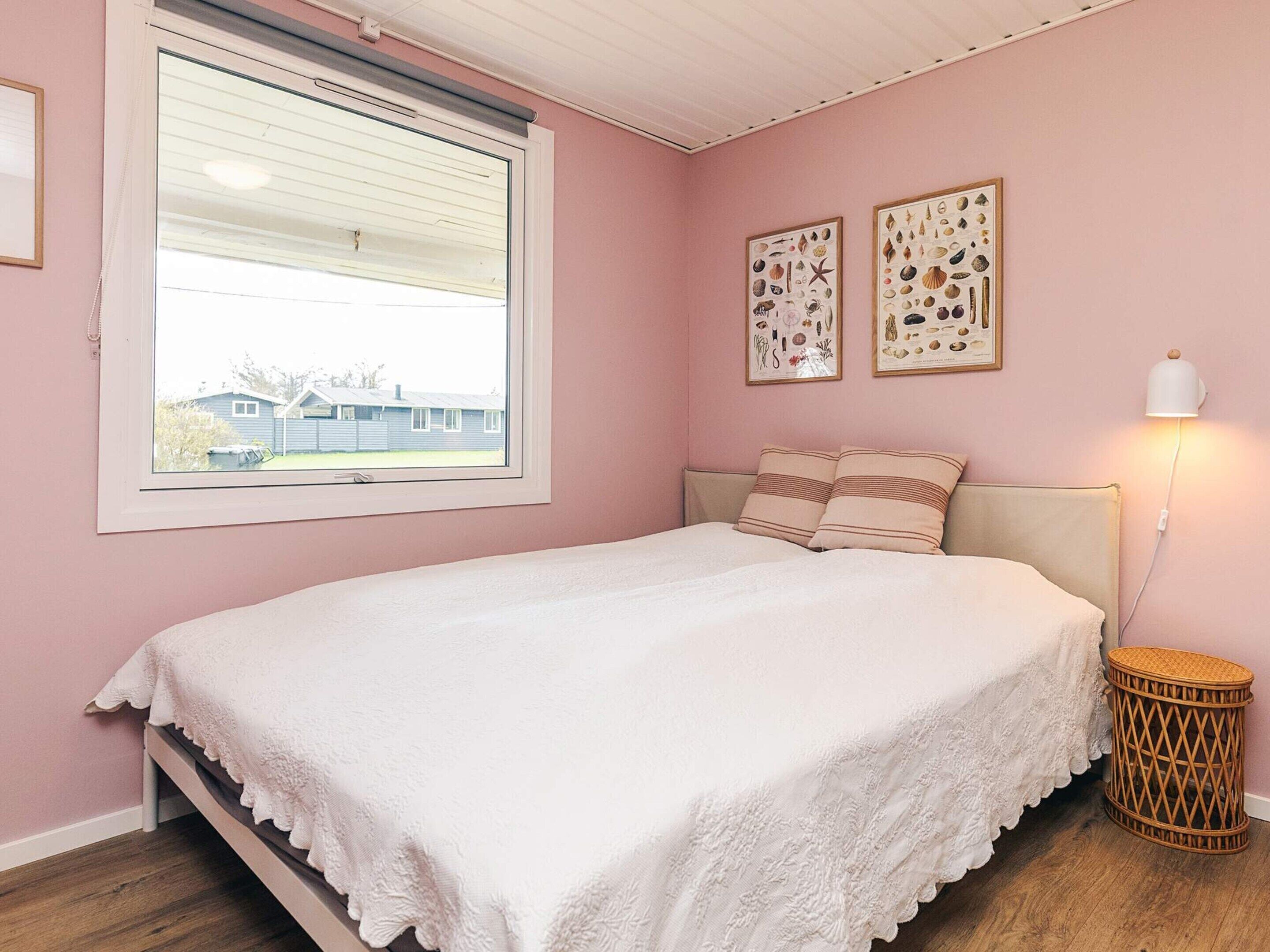 A bedroom with a bed, a window, two framed pictures, a bedside lamp, and a wicker side table.