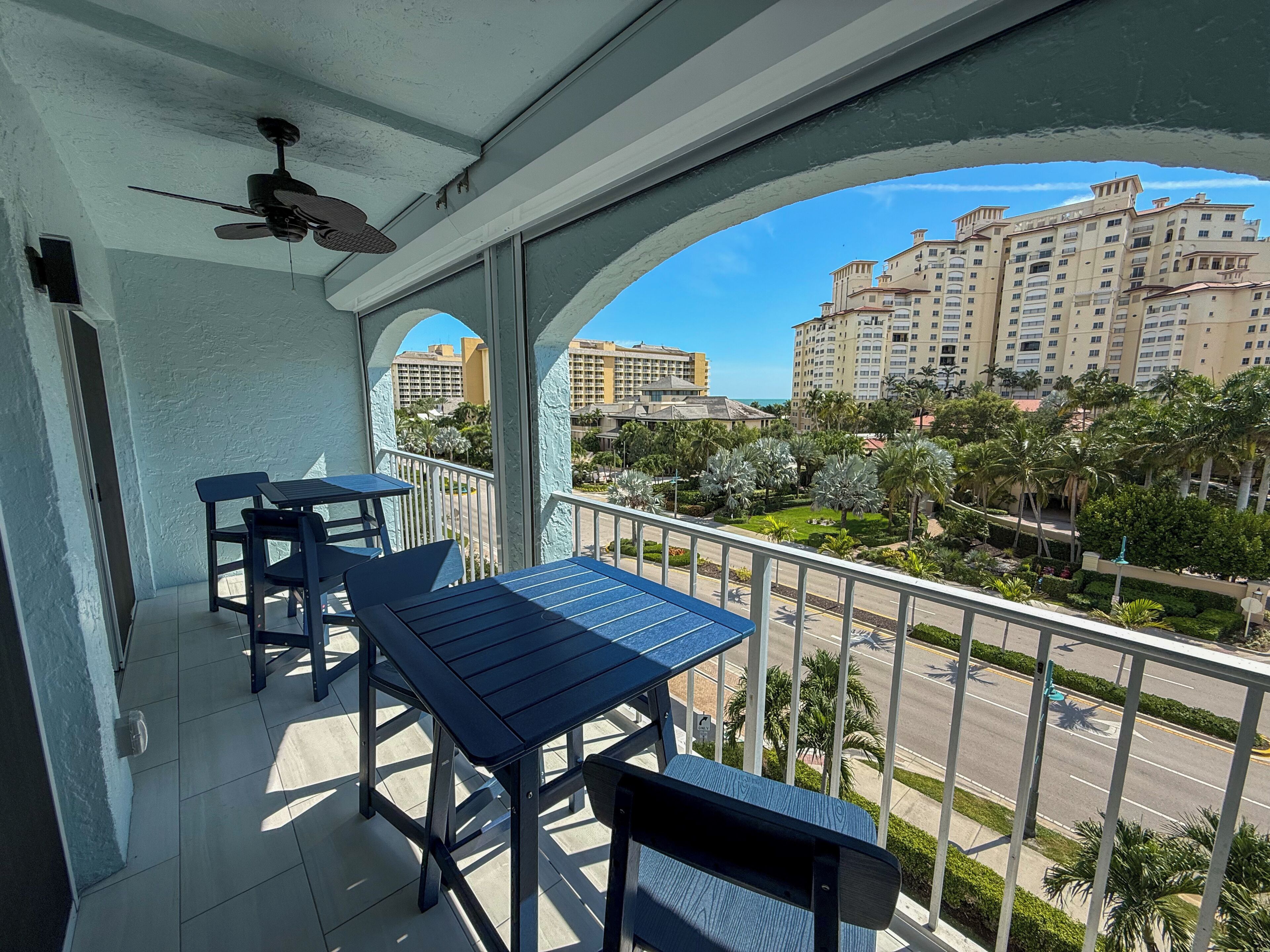 Een balkon met een tafel en stoelen, een plafondventilator en uitzicht op een stadsbeeld met gebouwen en palmbomen.