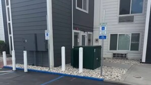 Un parking avec des places délimitées, des bollards et un bâtiment à la façade grise et aux fenêtres.