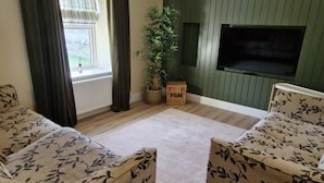 A living room with two patterned sofas, a wooden floor, a green wall, a ceiling fan, a window with curtains, and a plant. - Peaceful 2-bedroom house in DL perfect for relaxing getaways (DL)