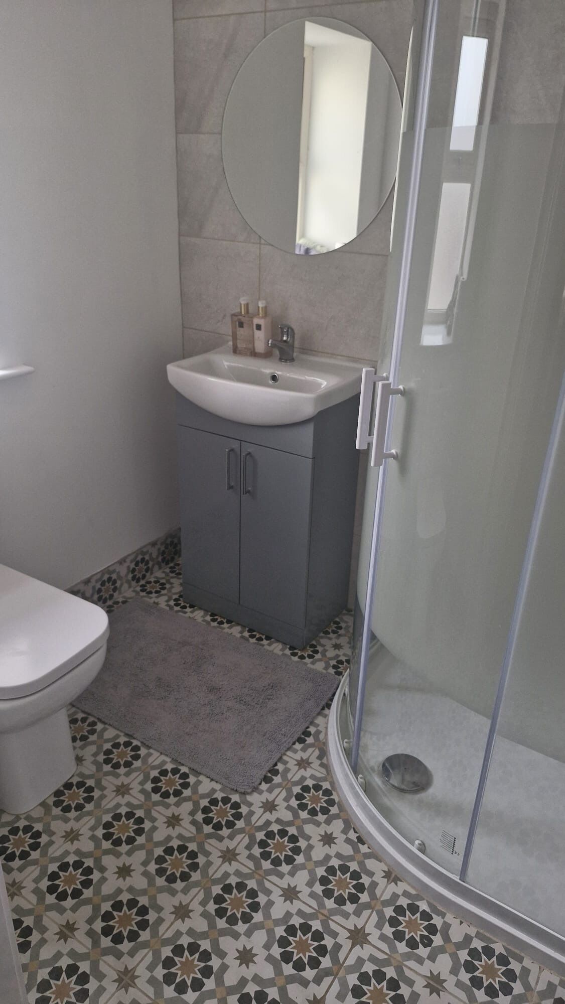 Et badeværelse med toilet, vask med spejl, brusebad og gulvfliser med mønster.
