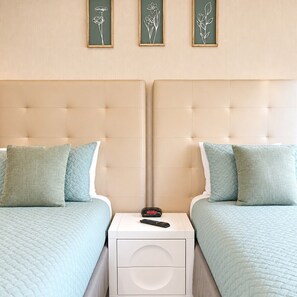 Een kamer met twee eenpersoonsbedden, elk met een kussen en een gewatteerde deken. Tussen de bedden staat een nachtkastje met een afstandsbediening.