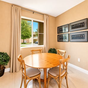 A dining room with a round wooden table, four chairs, beige curtains, a window with a view of trees and buildings, and framed wall art. - LuxuryResort~MagicVillage~Ensuite Baths~NearDisney (Kissimmee)