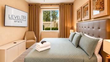 A hotel room with a bed, a desk, a chair, a ceiling fan, a window with curtains, and framed artwork on the wall.