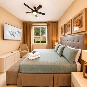 Habitación de hotel con cama, escritorio, silla, ventilador de techo, ventana con cortinas y cuadros en la pared.