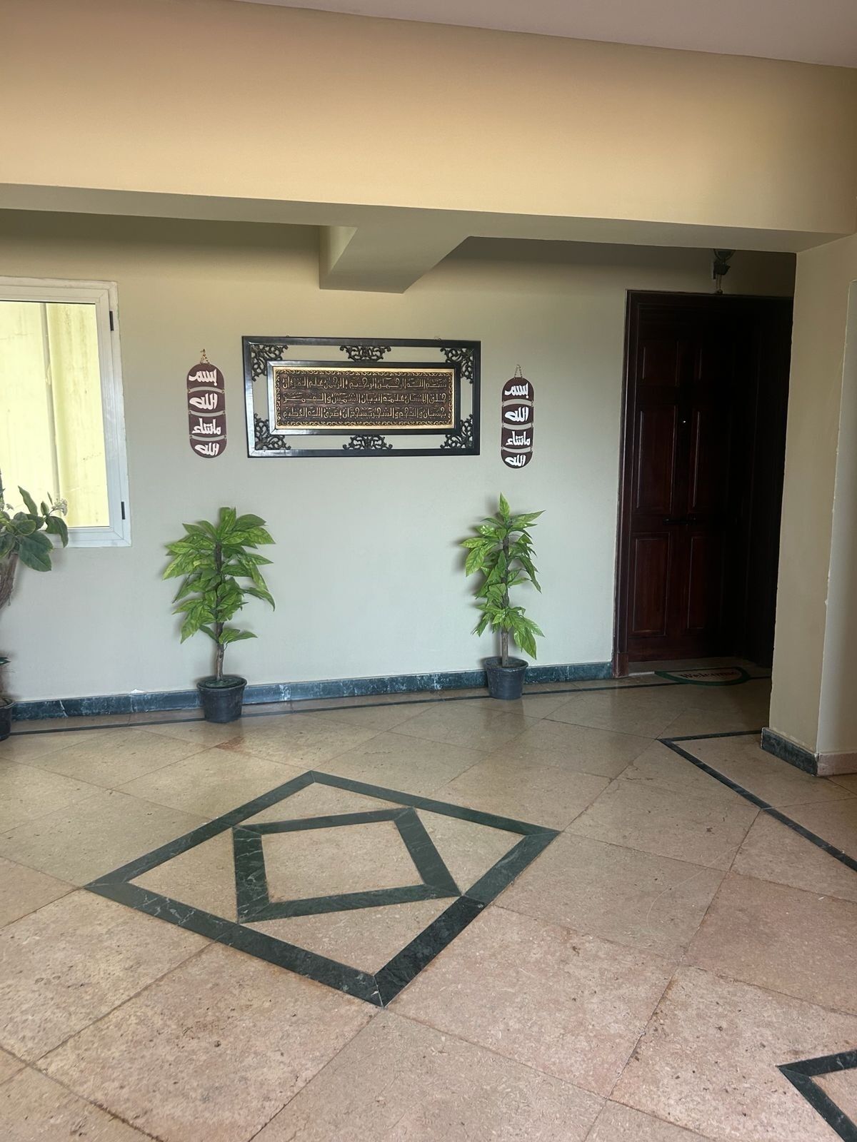 A hallway with a geometric tile pattern, two potted plants, a framed Arabic calligraphy piece, and a wooden door.