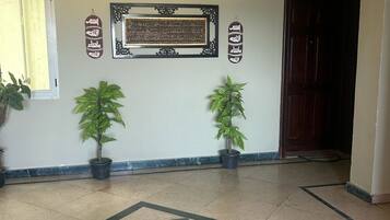 A hallway with a geometric tile pattern, two potted plants, a framed Arabic calligraphy piece, and a wooden door.