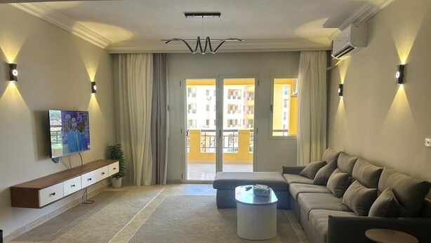 A modern living room with a sofa, coffee table, and TV on a wall. - Modern 2-bedroom Apt | Dreamland Golf & Pool View (Giza Governorate)