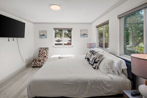 Un dormitorio con una cama grande, un televisor de pantalla plana en la pared, una ventana con vistas a árboles y una mesita con una lámpara.