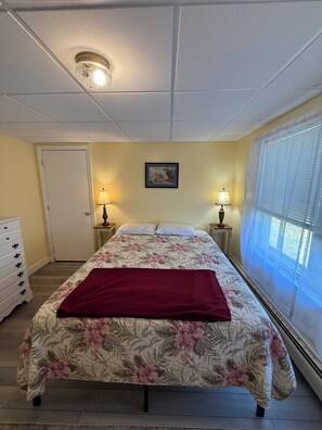 Een slaapkamer met een bed, twee bedlampen, een deur, een dressoir en een raam met jaloezieën.
