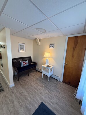 Une pièce avec un plancher en bois, un canapé bleu foncé orné d’un coussin à motif de homard, une table d  ’appoint blanche avec une lampe, et une porte en bois.