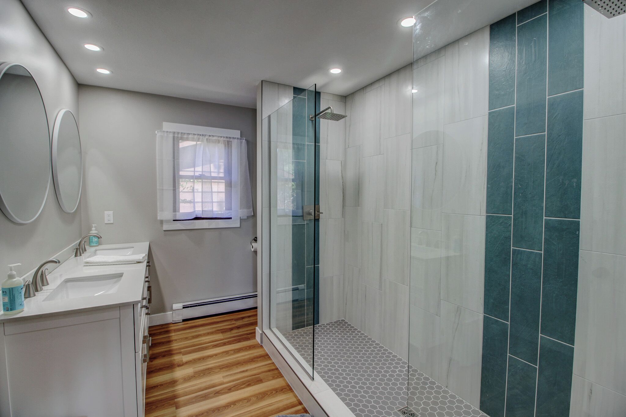 A modern bathroom with a glass shower enclosure, a sink with a mirror, and a window with a white curtain.