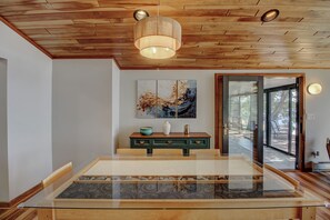 Un interior moderno con techo de madera, una mesa de vidrio con incrustación de piedra, una consola verde con objetos decorativos y un cuadro abstracto grande.