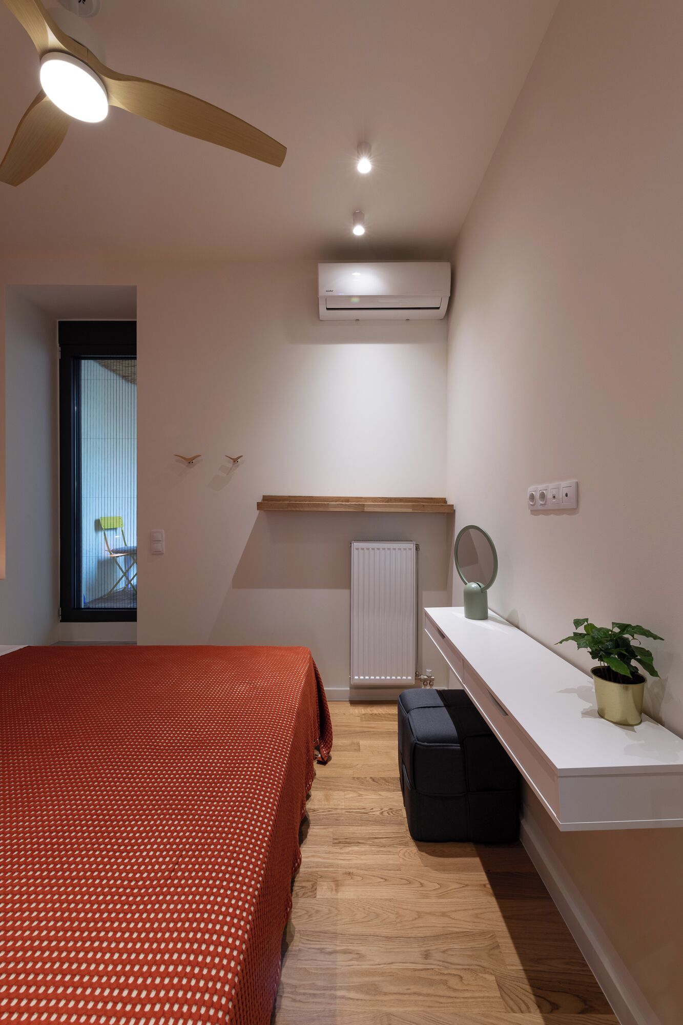 A bedroom with a bed, a ceiling fan, an air conditioner, a radiator, a small table with a mirror, a potted plant, and a window with blinds.