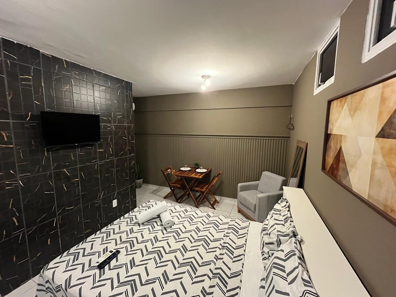 Quarto com cama, televisão na parede, mesa de jantar com cadeiras e uma poltrona cinza.