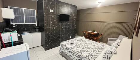 A compact hotel room with a bed, a small kitchenette, a seating area, and a TV.