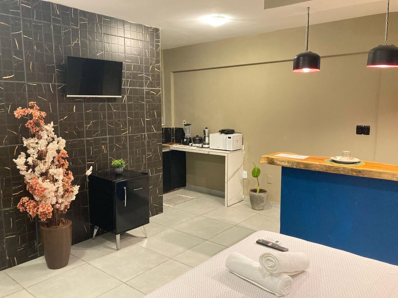 A modern hotel room with a black cabinet, a desk with a microwave, a TV on the wall, a reception desk, and a potted plant.