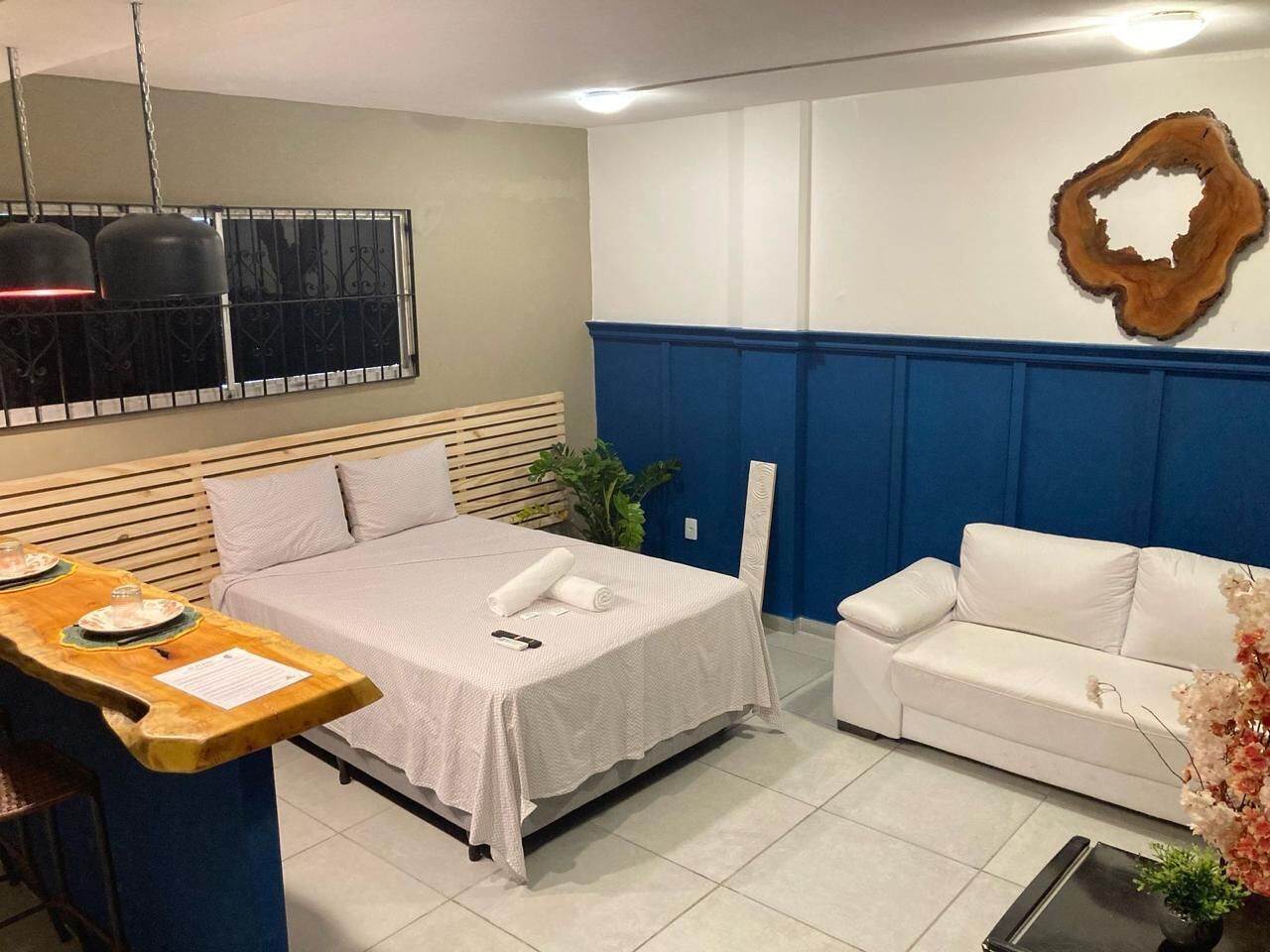 Habitación de hotel con cama, sofá blanco, mesita de madera, lámpara fijada a la pared y un adorno de madera.