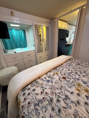 Un dormitorio con cama, tocador con espejo y una kitchenette al fondo.