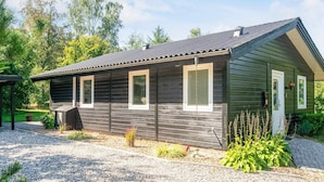 Ein eingeschossiges Holzhaus mit einem Schotterweg, umgeben von Grünflächen und Bäumen.