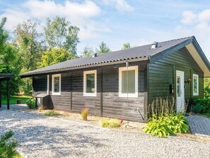 A single-story wooden cabin with a gravel driveway, surrounded by greenery and trees. - 4 Star Holiday Home in Farvang (Farvang)