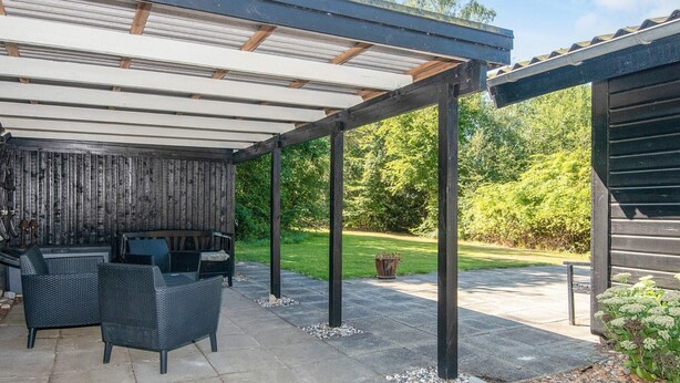 Ein möblierter Patio-Bereich im Freien, mit Überdachung, Steinboden und Blick auf einen üppigen Garten.