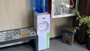 Un dispensador de agua, un rótulo rojo de emergencia, un cuadro enmarco, una planta en maceta y un banco de azulejos.