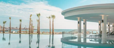 A pool area with palm trees, a glass-enclosed lounge, and a view of the sea.