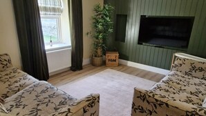 Una sala de estar con dos sofás estampados, piso de madera, pared verde, ventilador de techo, ventana con cortinas y una planta.