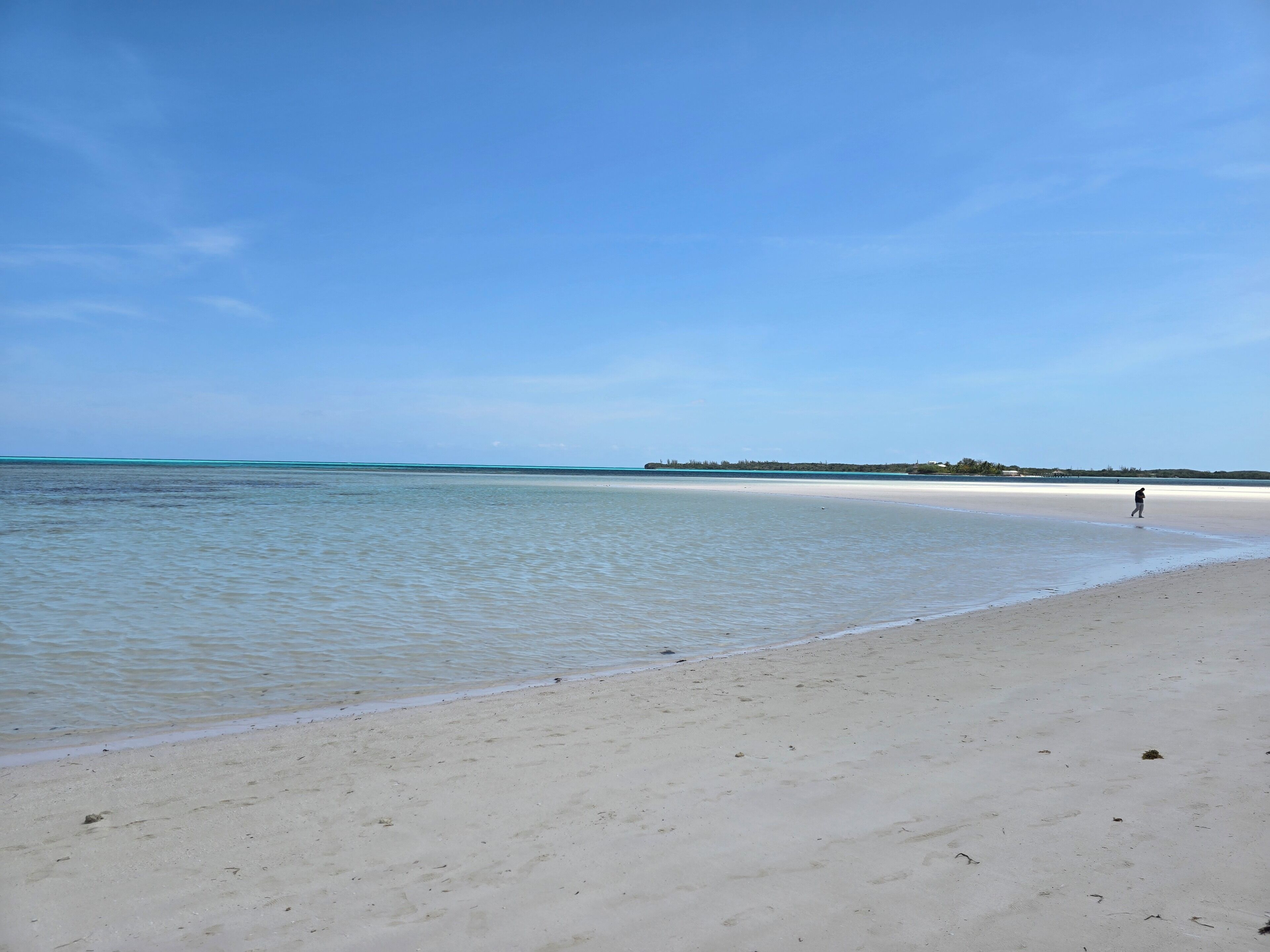 A beach with clear water, a person walking, and distant land.
