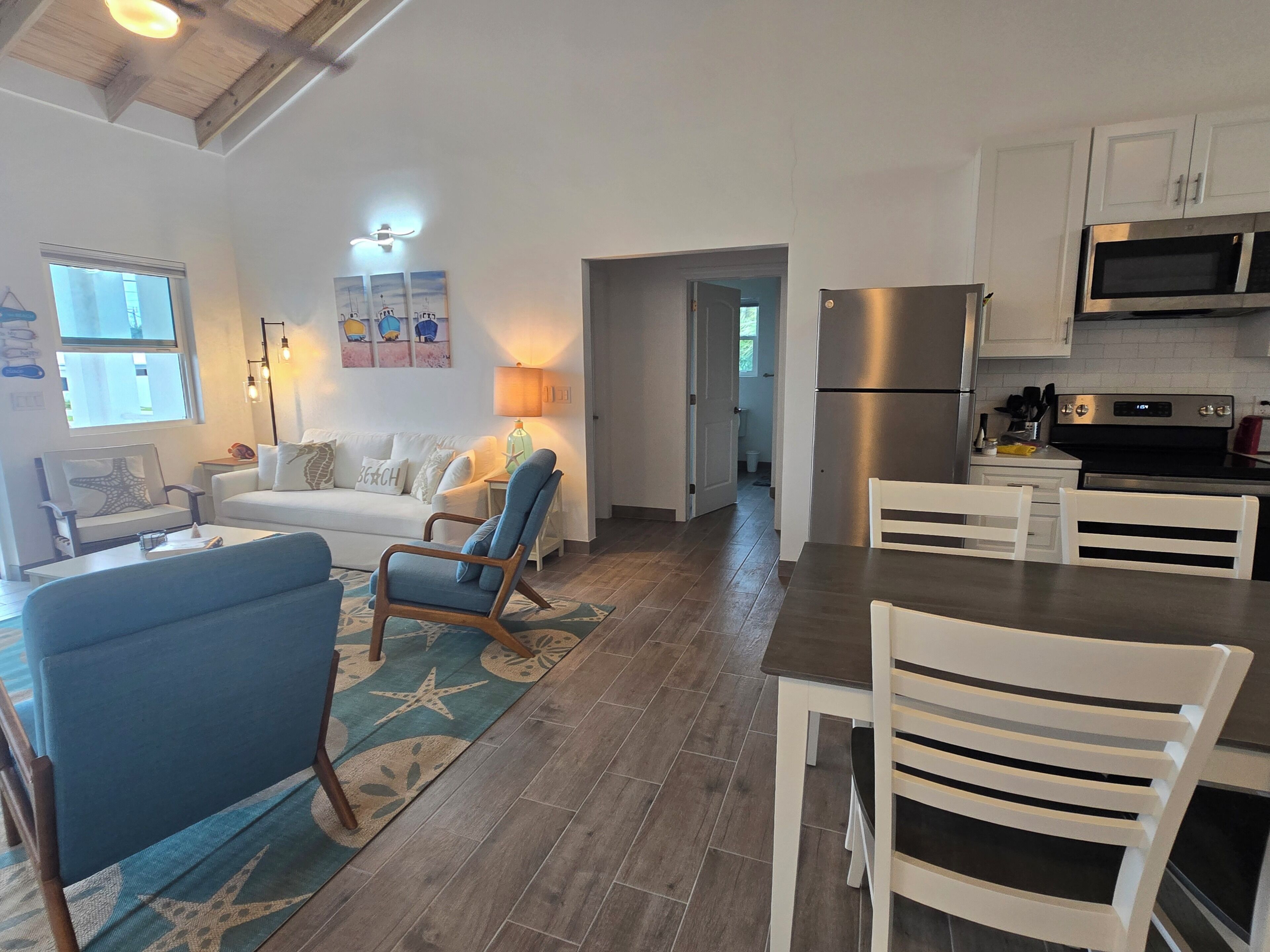 A modern kitchen with stainless steel appliances, a dining area with white chairs, and a living room with a sofa and blue armchairs.