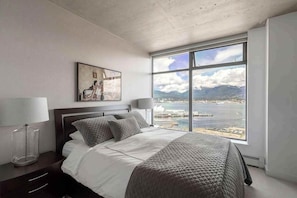 A modern bedroom with a large bed, a nightstand, a lamp, a framed picture, and a large window with a view of a body of water and mountains.
