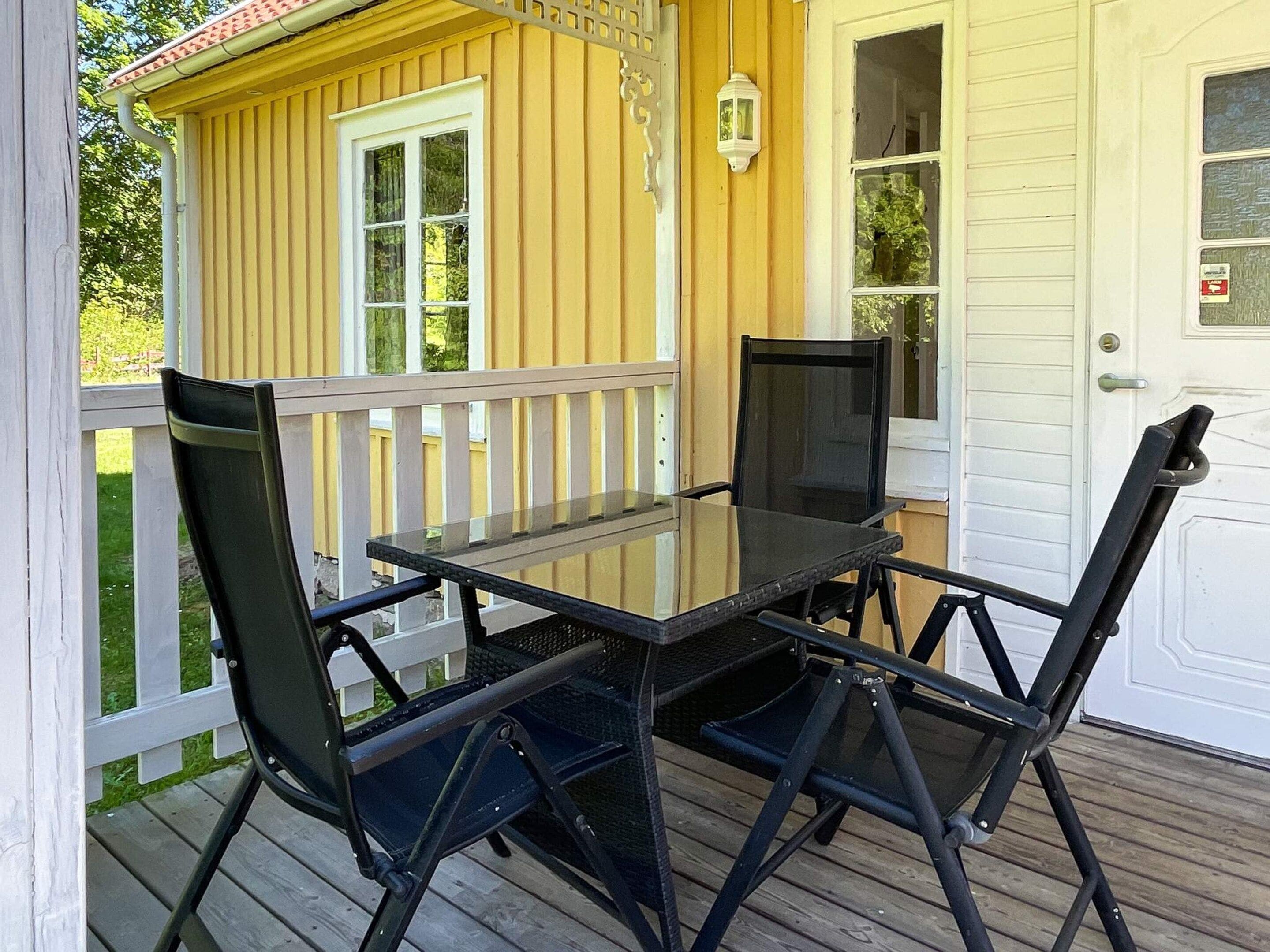 A glass-top table with four chairs on a wooden deck, a white door, and a yellow building with a white railing in the background.
