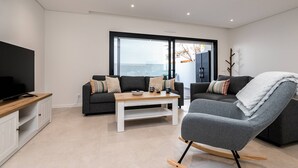 A modern living room with a sofa, a rocking chair, a coffee table, a TV, and a large window. - Concei Sol Modern Apartment (Tavira)