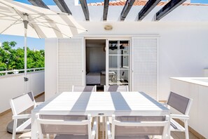 Eine Terrasse mit Tisch und Stühlen, eine Schiebetür, die zu einem Raum führt, und ein Sonnenschirm.