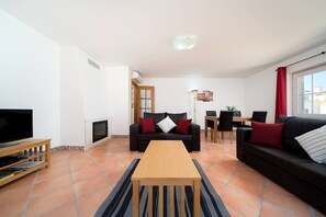 Ein Wohnzimmer mit Fernseher, Sofa, Couchtisch, Kamin, Essbereich und einem Fenster mit Vorhängen.