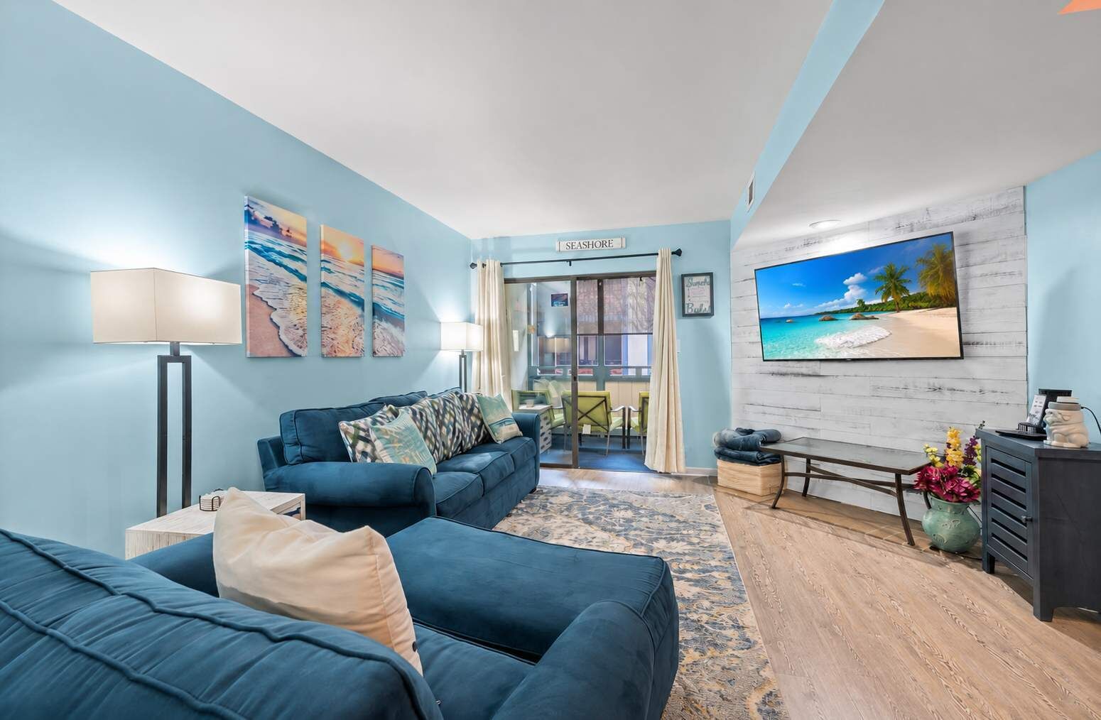 Un salon avec un canapé bleu, une lampe, un téléviseur et un tableau représentant une scène de plage.