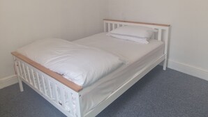 Ein weißes Bett mit Holz-Kopfstück in einem Zimmer mit schlichten weißen Wänden und einem grauen Teppich.