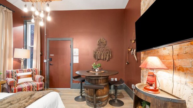 A cozy room with a bed, a patterned sofa, a round wooden table with chairs, a decorative wall piece, and a brick wall with a television. - StayRodeo | Republic | Red (Cincinnati)