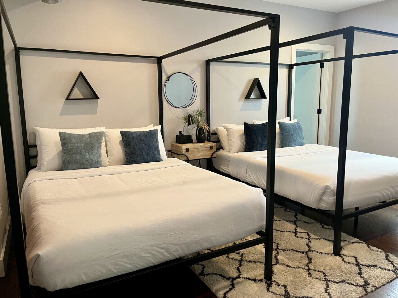 A four-poster bed with white bedding and blue pillows, a round mirror, and a small wooden side table with a plant.