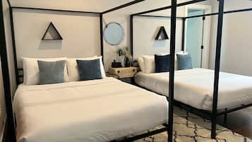 A four-poster bed with white bedding and blue pillows, a round mirror, and a small wooden side table with a plant.
