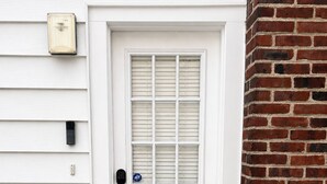 Uma porta branca com janela, um tapete de entrada com a palavra 'hello' e uma parede de tijolos à direita.