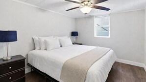 A bedroom with a bed, a ceiling fan, a nightstand, a lamp, and a window. - Leavenworth Apartment (Omaha)